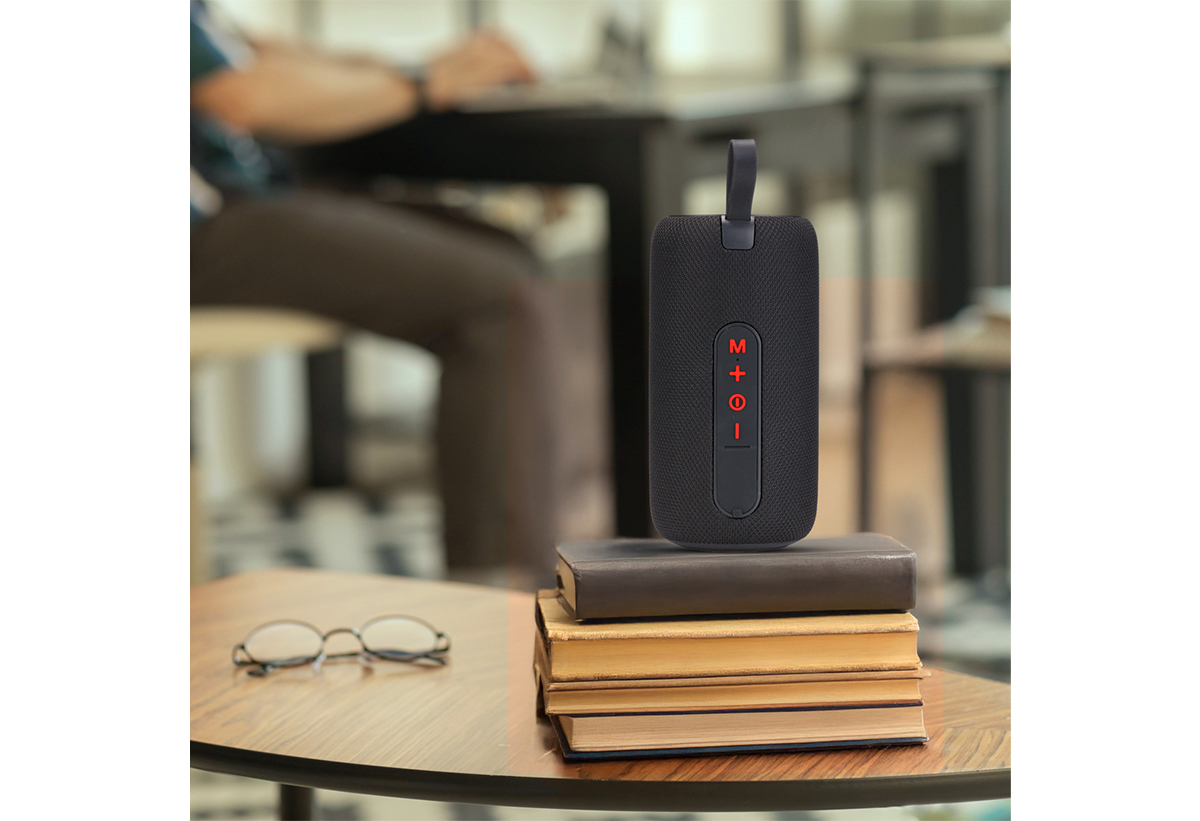 Απεικονίζεται το ηχείο πάνω σε στοίβα με βιβλία σε τραπέζι, δίπλα σε γυαλιά οράσεως.